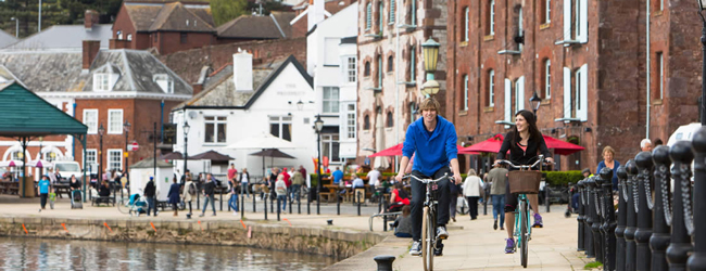Fahrradausflug entlang des Flusses in Exeter Zwei personen fahren Fahhrad am Ufer des Flusses in Exeter im Hintergrund Menschen und Cafes