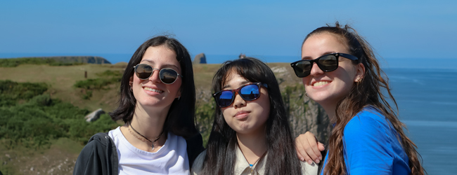 Drei Mädchen mit Sonnenbrillen stehen in Wales an der Küste vor dem Meer